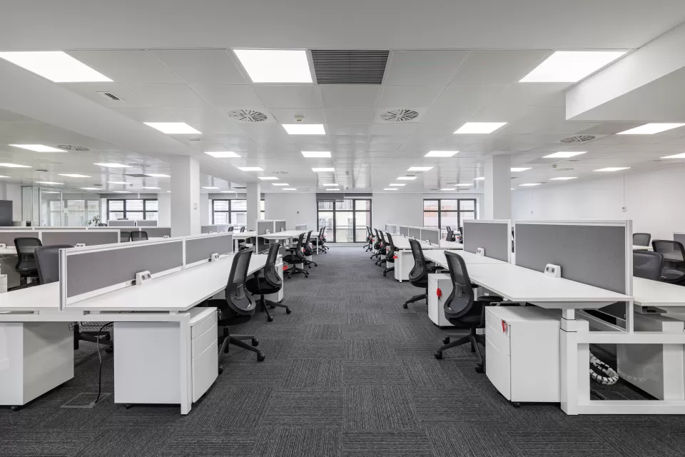 Large empty open-plan office with rows of white desks and black chairs under bright fluorescent lighting
